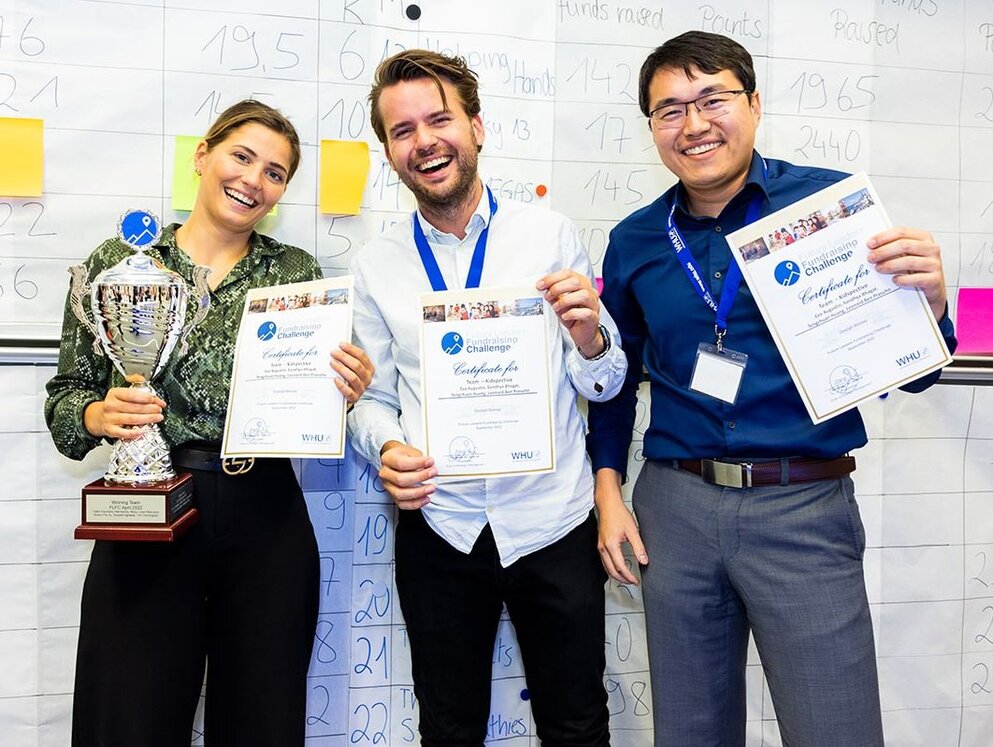Three smiling MBA students are holding up their Future Leaders Fundraising Challenge certificates. The young woman on the left is holiding a silver trophy.