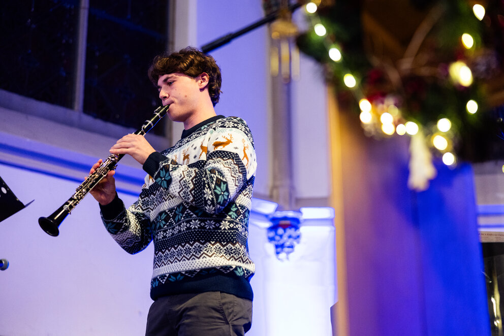Ein dunkelhaariger junger Mann in einem marineblau-weißen Weihnachtspullover im nordischen Stil spielt Klarinette in der Kapelle auf dem WHU Campus Vallendar.