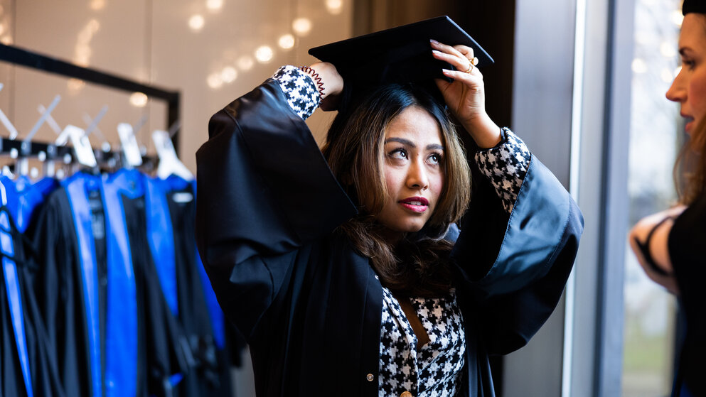 A dark-haired young woman stands in front of a rail of black gowns with blue sashes and puts a black graduation cap on her head.