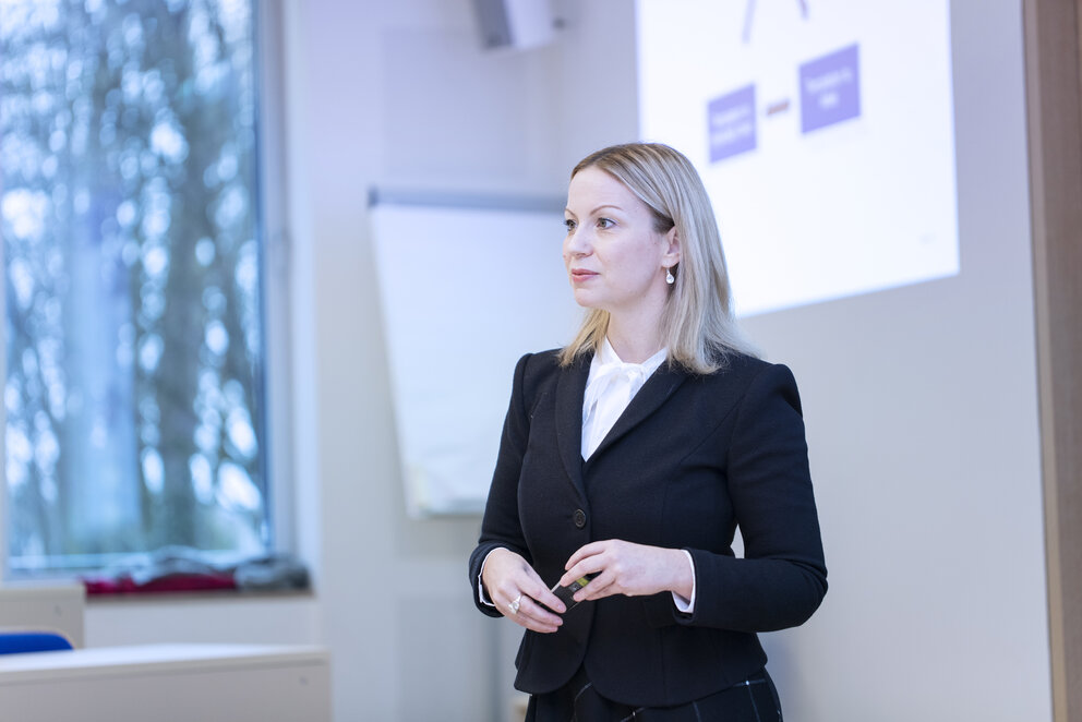 A woman with shoulder-length blonde hair wearing a black suit over a white blouse gives a presentation in a lecture room.