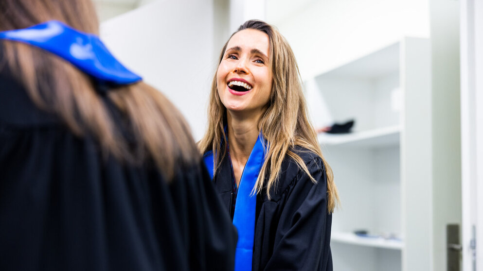 Eine junge Absolventin mit langem hellbraunem Haar, die einen schwarzen Talar mit einer blauen Schärpe trägt, lächelt breit in die Kamera.