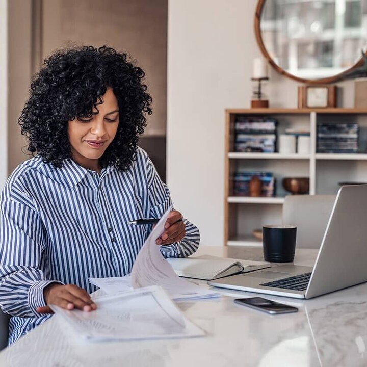 Eine junge Frau mit langem, schwarzem, lockigem Haar und blau-weiß gestreiftem Hemd arbeitet in ihrem Homeoffice vor ihrem Laptop und liest einige Papiere. 