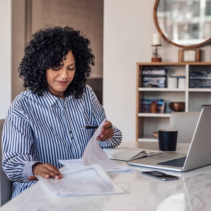 A young woman with long black curly hair, wearing a blue and white striped shirt, works in front of her laptop in home office and reads some papers.