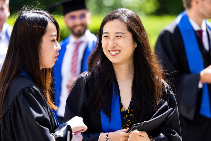 Two female graduates wearing black gowns with blue sashes chat to each other in the sunshine.