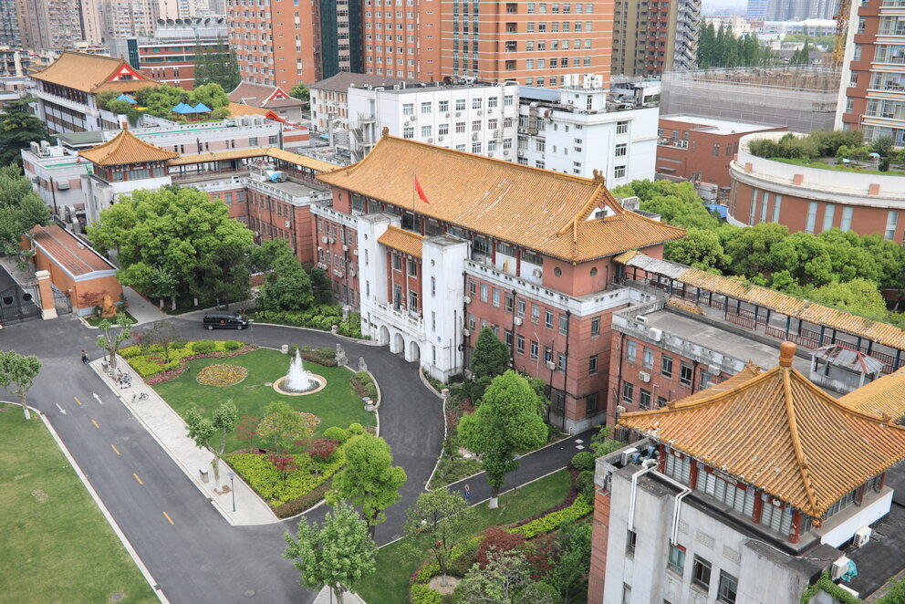 A birds-eye view of red-brick buildings with curving oriental roofs around a semi-circular park with ornamental shrubs and a fountain.