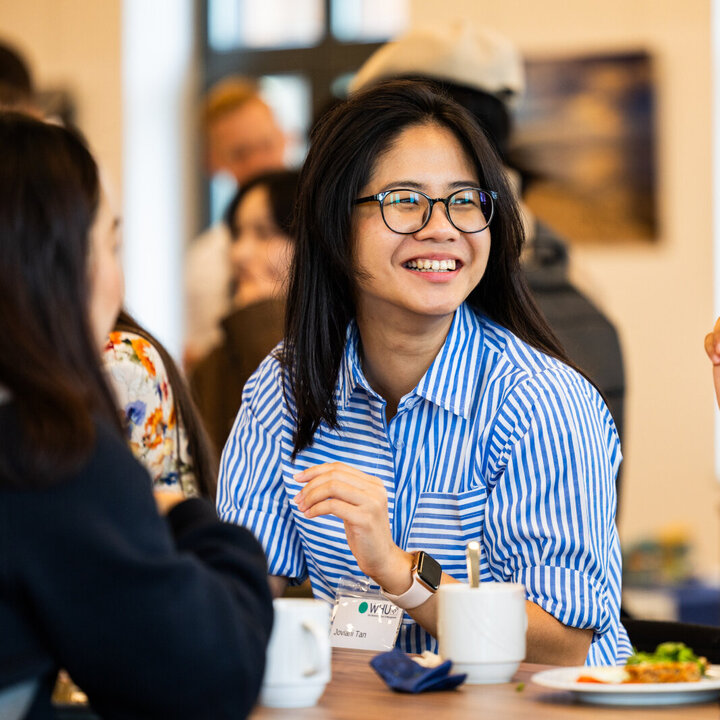 Eine junge ostasiatische Frau mit einer dunkel gerahmten Brille und einem blau-weiß gestreiften Hemd lächelt die Frau neben ihr an, während sie essen.
