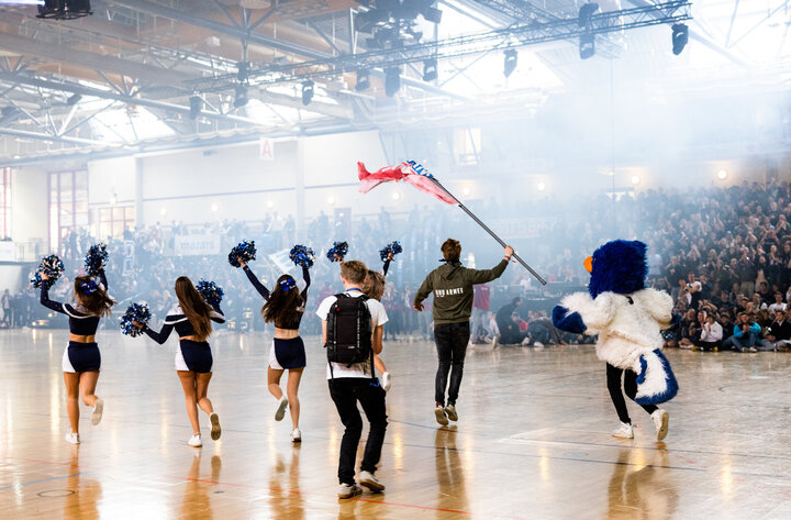 Young women dressed as cheerleaders and a young man carrying a colourul flag celebrate the Euromasters tournament in front of crowd of spectators in a sports hall. The WHUhu, dressed in a blue and white owl costume, is running beside them.