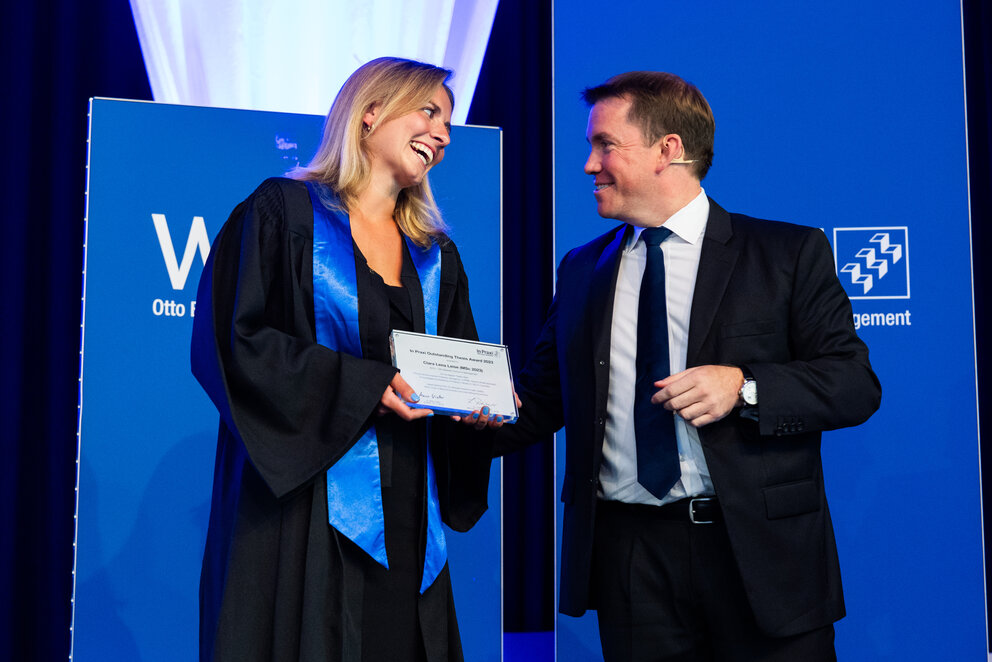 A smiling Master Student with shoulder-length blonde hair and wearing a black gown with a blue sash, receives the In Praxi Outstanding Thesis Award from a short-haired man wearing a dark suit and tie over a white shirt.