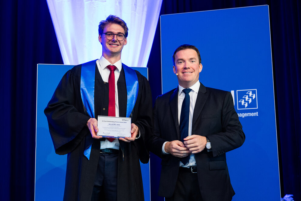 A slim young man with dark hair and glasses, wearing a graduation gown with a blue sash, stands next to a shorter man in a dark suit with a white shirt and a navy blue tie. They are both smiling at the camera, and the graduate proudly holds a certificate in his hand.