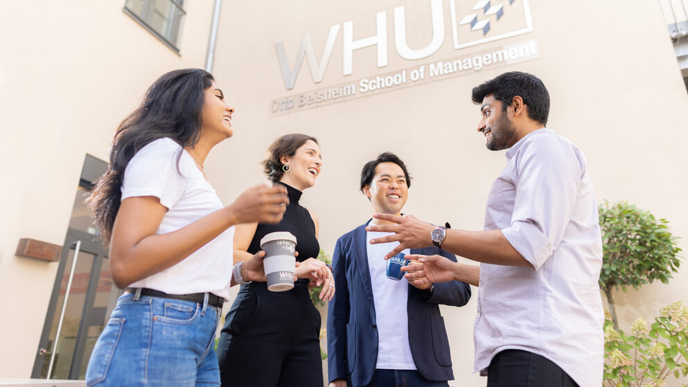 Four young people of mixed nationalities stand outside chatting and drinking coffee during a break on WHU Campus Düsseldorf.