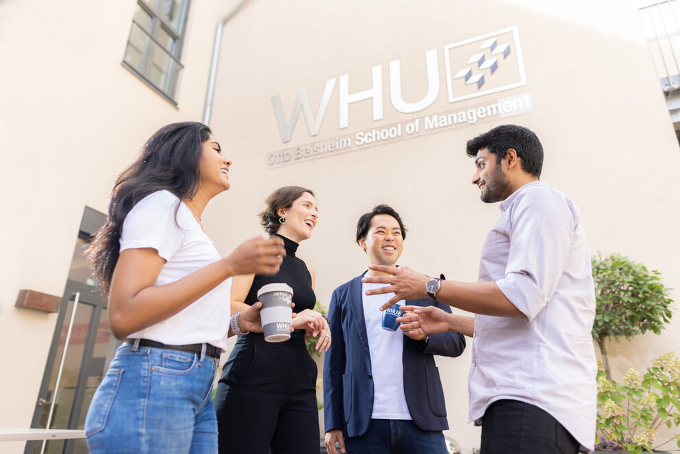 Vier junge Leute gemischter Nationalität stehen während einer Pause auf dem WHU-Campus Düsseldorf im Freien, unterhalten sich und trinken Kaffee