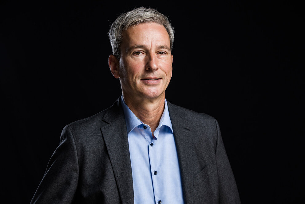 A grey-haired man wearing a blue shirt and dark suit jacket looks into the camera
