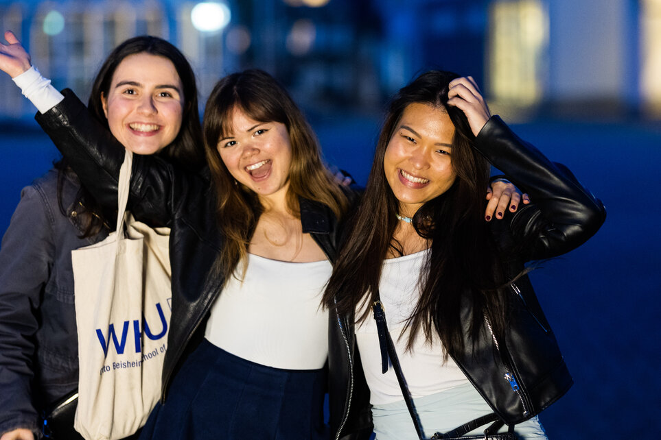 Three young women with dark hair pose, laughing into the camera, at an event for Integration@Vallendar. The woman in the middle has a white cloth bag emblazoned with the WHU logo hooked over her right arm.